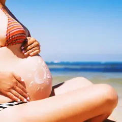 Brzuszek w ciąży na plaży, posmarowany kremem z filtrem w kształcie słońca. Bezpieczne opalanie w ciąży to podstawa.