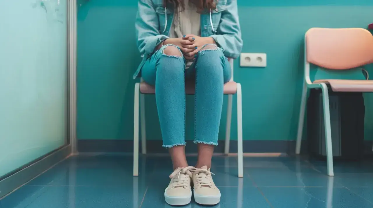 Dziewczyna w podartych dżinsach siedzi na krześle, czekając na wizytę. Jej dłonie splecione na kolanach mogą sugerować niepokój lub pochwicę.