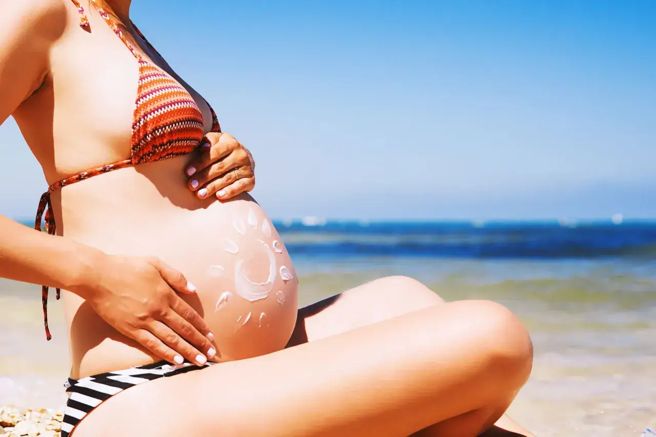 Brzuszek w bikini na plaży, z kremem przeciwsłonecznym w kształcie słońca. Bezpieczne opalanie w ciąży to podstawa.